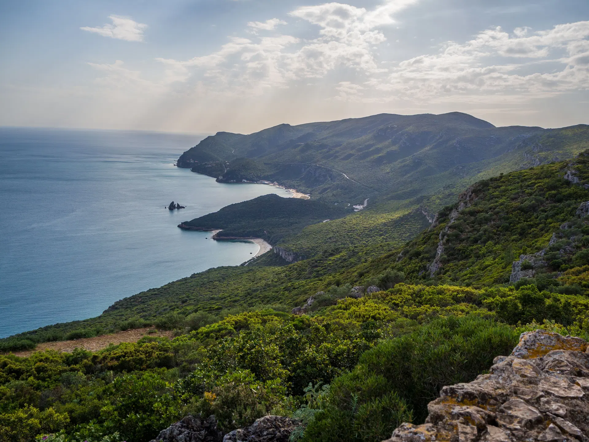 Scenic coastal hillside landscape with green mountains, sea views and small bays under a cloudy sky.
