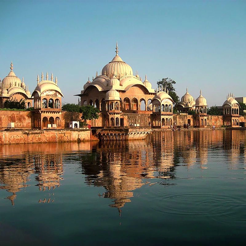 Riverside temple complex with domes reflected in calm water under a clear blue sky.